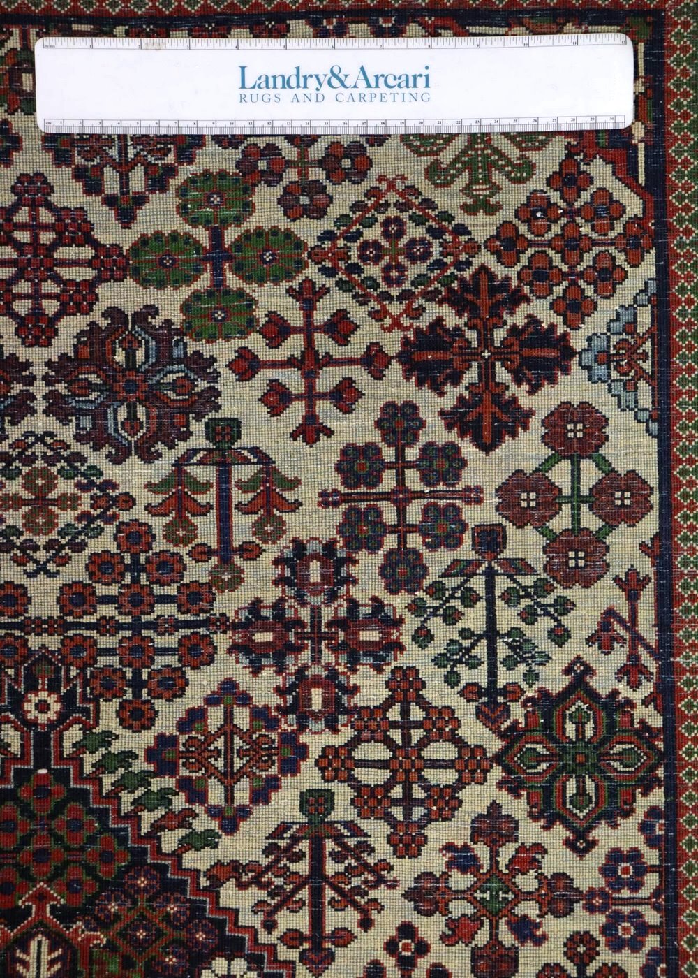 Close-up detail of antique tribal rug with intricate floral and geometric patterns in red, blue, and green on an off-white background.