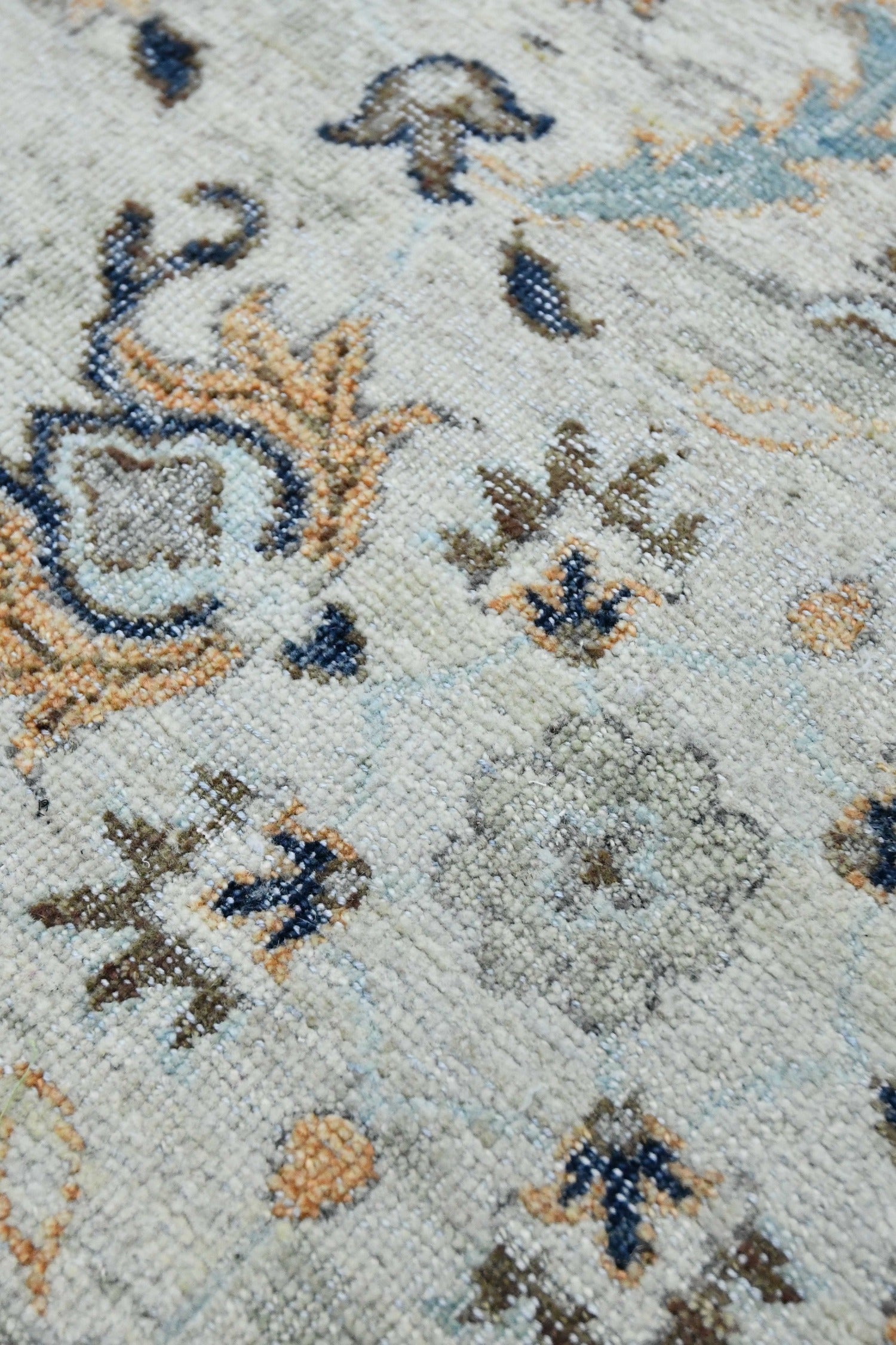 Close-up of handwoven rug detail, showcasing a light beige base with muted blue, orange, and brown floral patterns.
