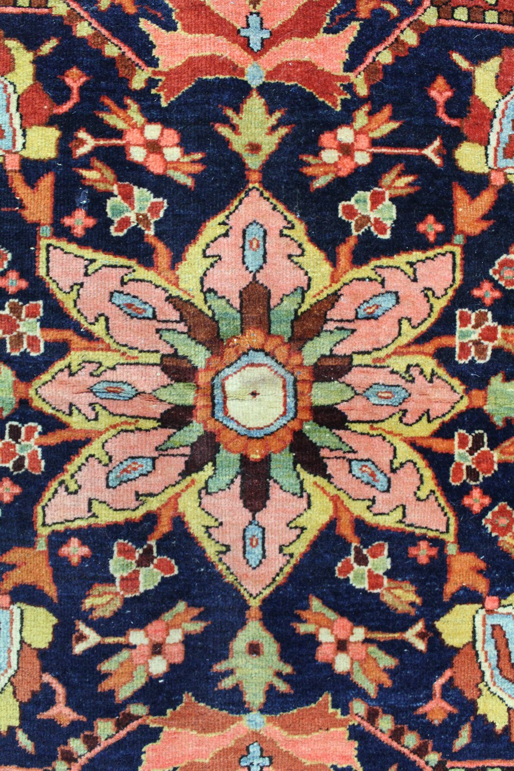 Close-up of a richly colored antique rug, showcasing intricate floral details in pink, teal, and gold against a dark blue background.
