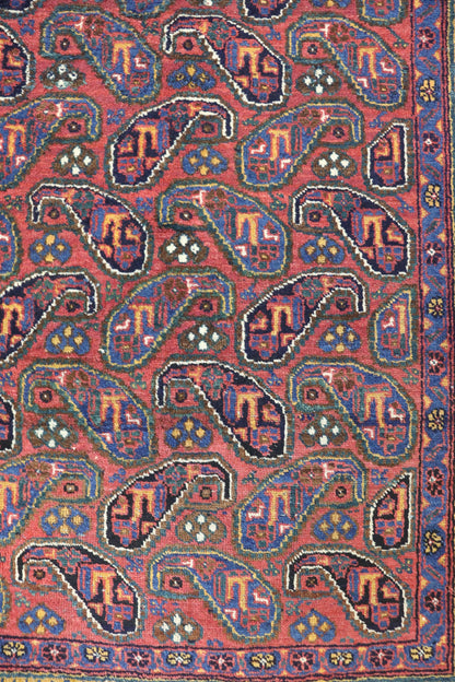 Handwoven tribal rug detail: red base with intricate paisley pattern in navy, teal, and gold.
