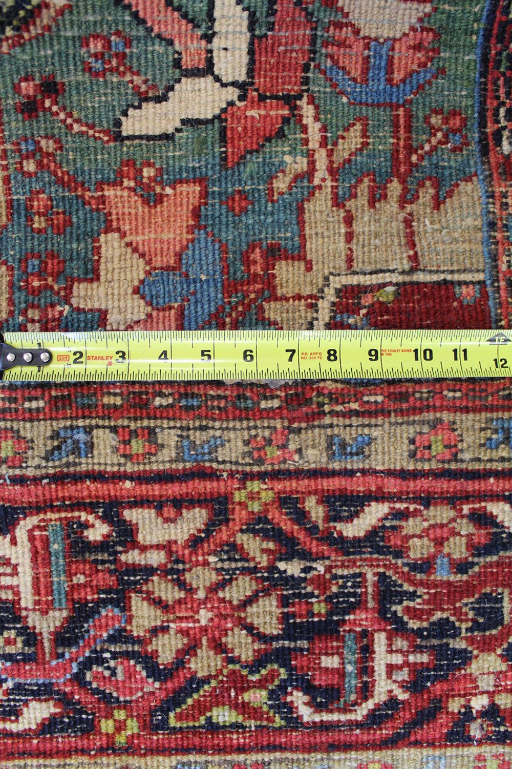 Close-up of antique Heriz rug detail, showing intricate floral pattern in red, blue, and beige, measured with tape.
